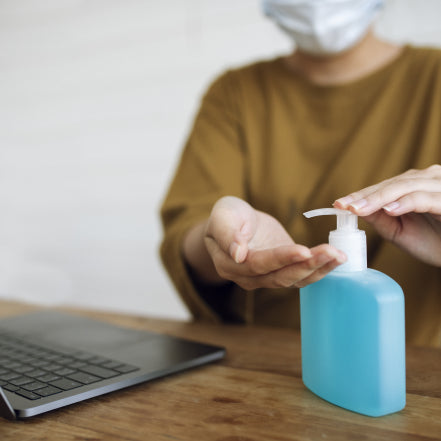 Women with face mask on sanitizing her hands while sat at desk on laptop